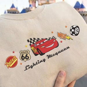 May include: Beige crew neck sweatshirt with a red Lightning McQueen graphic and the text "Lightning McQueen". The design includes a checkered flag, Route 66 logo, and a tyre. The sweatshirt has a classic, casual style.