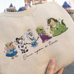 May include: Beige sweatshirt featuring embroidered cartoon characters, including a fairy, pig, snowman, and frog, with the text "Once upon a time." The characters are in various colors, and the sweatshirt has a crew neck.