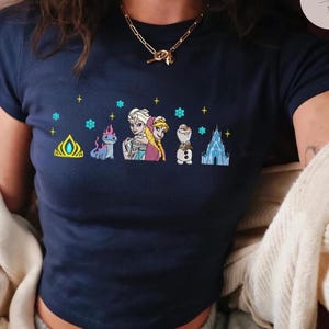May include: Navy blue crop top featuring a colorful embroidered design of characters and elements from a popular animated movie. The design includes a crown, a dragon, two princesses, a snowman, and an ice castle, with snowflakes and stars.