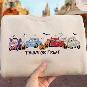 May include: A beige crewneck sweatshirt featuring an embroidered design of four cartoon cars dressed in Halloween costumes, including a vampire, a devil, and a mummy, alongside orange pumpkins, ghosts, bats, and the text TRUNK OR TREAT.