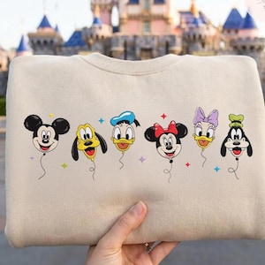 May include: Beige crewneck sweatshirt featuring embroidered Disney characters as balloons. Mickey Mouse, Pluto, Donald Duck, Minnie Mouse, Daisy Duck, and Goofy are depicted with smiling faces and colorful accents. A fun, whimsical design.