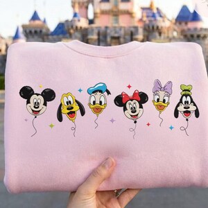 May include: A light pink sweatshirt featuring a graphic of Mickey Mouse, Pluto, Donald Duck, Minnie Mouse, Daisy Duck, and Goofy as balloon characters. Each character is depicted with a smiling face and attached to a string. The background shows a castle.