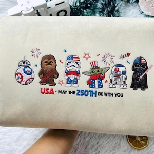 Op de afbeelding: Een cr&egrave;mekleurig kussen met geborduurde Star Wars-personages in patriottische kleding. Het ontwerp bevat BB-8, Chewbacca, een Stormtrooper, Grogu, R2-D2 en Darth Vader, met de tekst "USA - May the 250th be with you."
