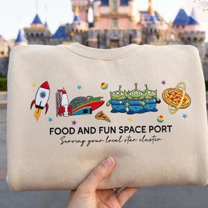 May include: Beige sweatshirt with a colorful graphic featuring a rocket, space port, pizza, and three green aliens. The text reads "FOOD AND FUN SPACE PORT" and "Serving your local star cluster."