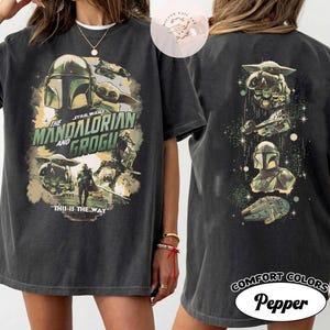 May include: Dark grey t-shirt featuring a Star Wars design with The Mandalorian and Grogu. The front graphic includes the text "This is the Way." The back of the shirt has additional Star Wars imagery. The shirt is labelled "Comfort Colors Pepper."