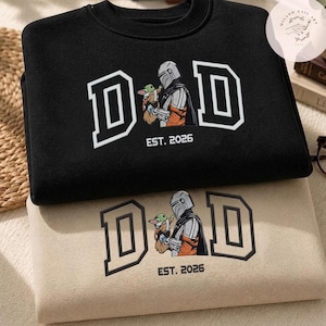 May include: Two sweatshirts, one black and one beige, are stacked. Both feature the word "DAD" in a white outlined font, with an image of a Mandalorian character holding a small green creature. The text "EST. 2026" is below the image.