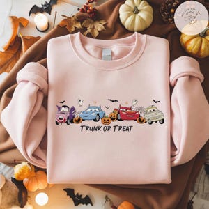 May include: A light pink crewneck sweatshirt featuring a graphic of four cartoon cars dressed in Halloween costumes, including a vampire, a mummy, and others, surrounded by pumpkins, bats, and ghosts, with the text TRUNK OR TREAT printed below.