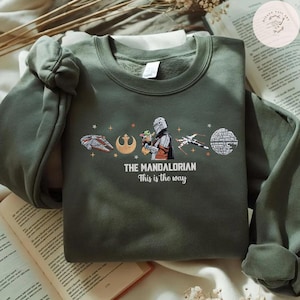 May include: Olive green sweatshirt featuring a graphic from "The Mandalorian." The design includes the text "THE MANDALORIAN" and "This is the way," along with images of the Millennium Falcon, a Rebel symbol, the Mandalorian, Grogu, and other Star Wars elements.