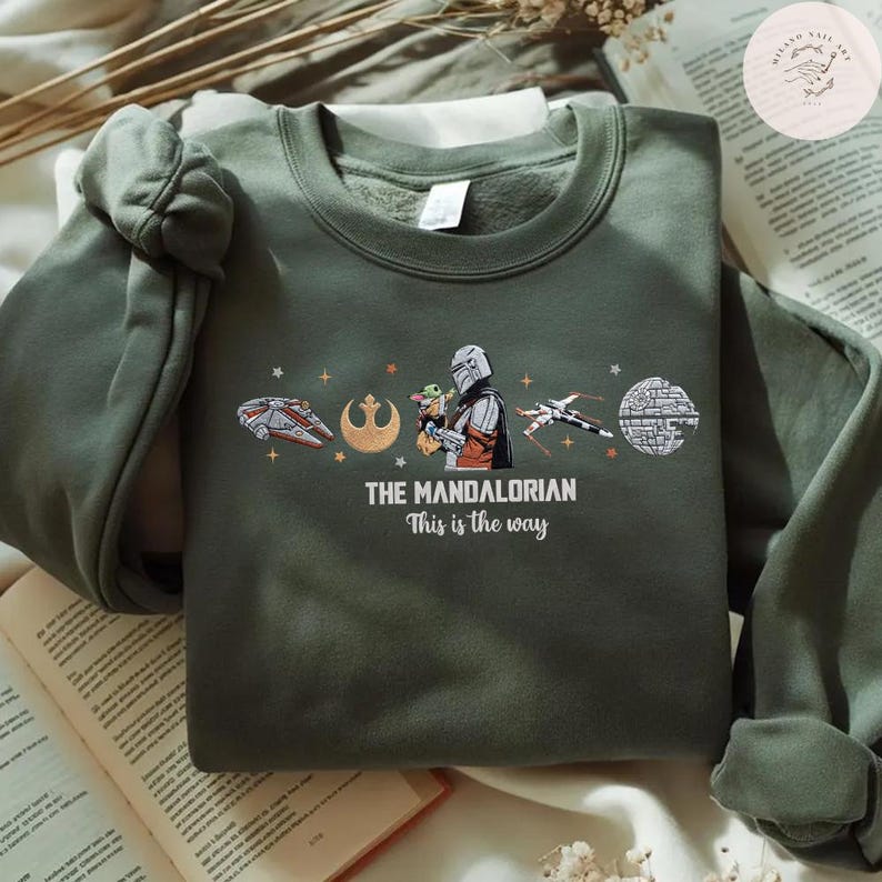 May include: Olive green sweatshirt featuring a graphic from "The Mandalorian." The design includes the text "THE MANDALORIAN" and "This is the way," along with images of the Millennium Falcon, a Rebel symbol, the Mandalorian, Grogu, and other Star Wars elements.