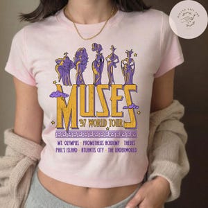 May include: A light pink t-shirt featuring a graphic of the Muses from Hercules. The design includes the word "MUSES" in yellow and purple, with "'97 WORLD TOUR" below. Additional text lists locations like Mt. Olympus and Thebes.