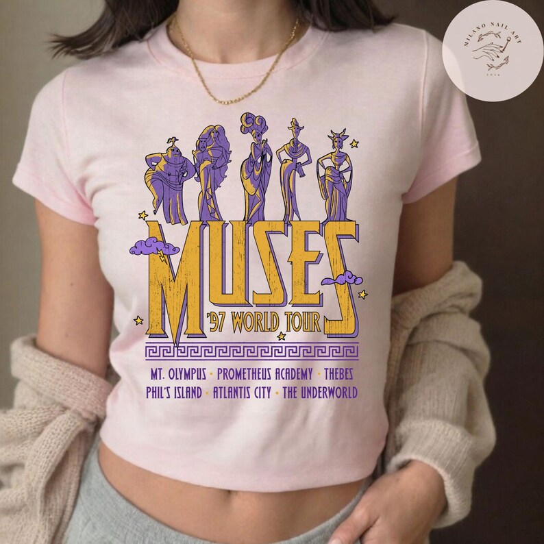 May include: A light pink t-shirt featuring a graphic of the Muses from Hercules. The design includes the word "MUSES" in yellow and purple, with "'97 WORLD TOUR" below. Additional text lists locations like Mt. Olympus and Thebes.