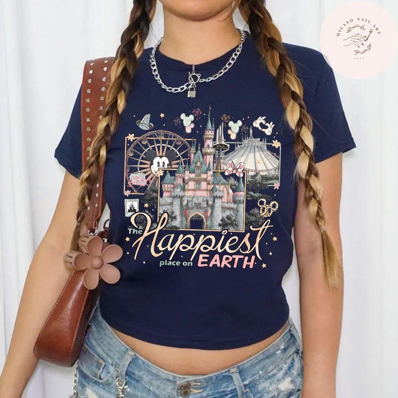May include: Navy blue t-shirt featuring a colorful graphic of a castle, fireworks, and a Ferris wheel. The text "The Happiest Place on Earth" is displayed below the graphic. The shirt has short sleeves and a crew neck.