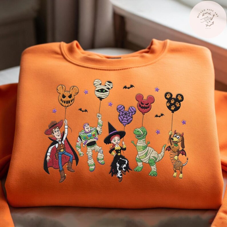 May include: An orange crewneck sweatshirt featuring an embroidered design of five animated characters dressed in Halloween costumes, each holding a Mickey Mouse-shaped balloon. The characters include a cowboy, a space ranger, a cowgirl, a dinosaur, and a dog.