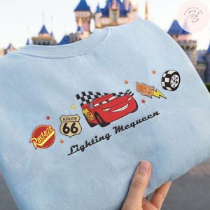 May include: Pale blue sweatshirt with a graphic of Lightning McQueen from the Cars film. The design includes Route 66, a checkered flag, and the text "Lightning McQueen". The sweatshirt has a crew neck.