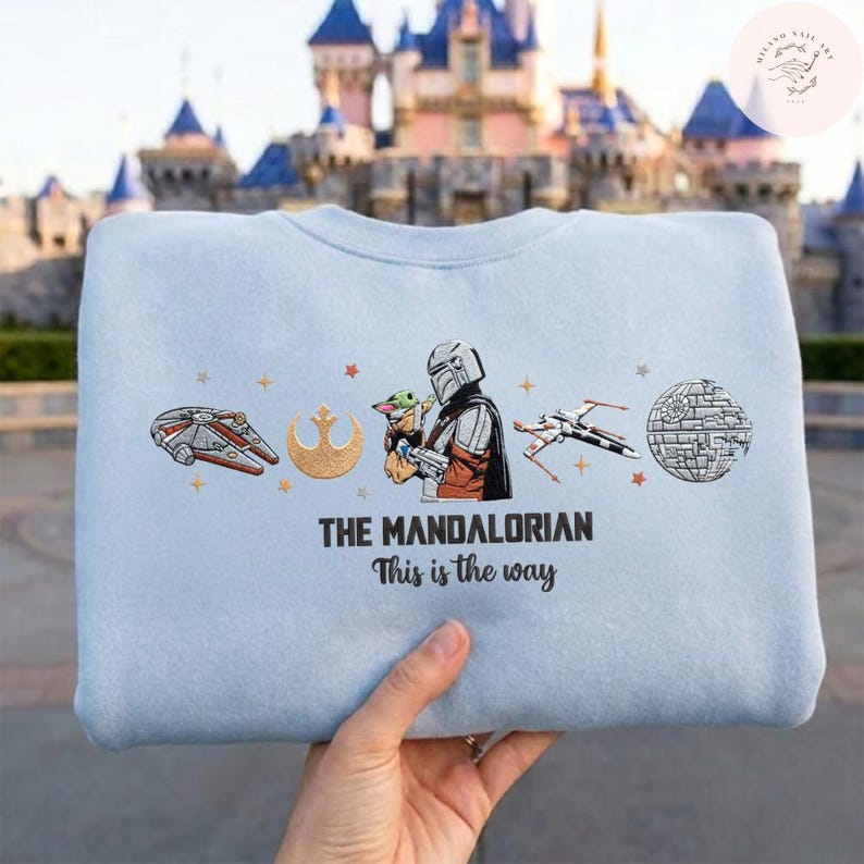May include: Light blue crewneck sweatshirt featuring embroidered images of Star Wars characters and symbols. The design includes the Millennium Falcon, a Rebel Alliance symbol, The Mandalorian with Grogu, an X-wing, and the Death Star. Text reads "THE MANDALORIAN This is the way."