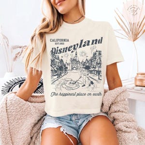 May include: Cream-colored t-shirt featuring a Disneyland graphic. The design includes the text "CALIFORNIA EST.1955 Disneyland" above a detailed illustration of a park scene with fireworks and Mickey Mouse. The phrase "The happiest place on earth" is printed below.