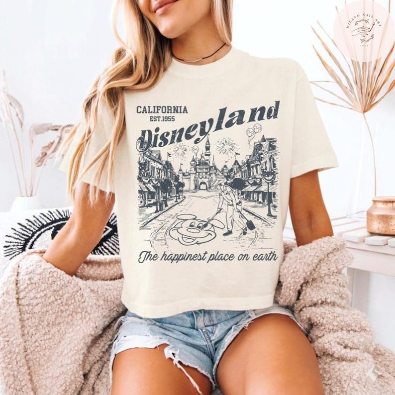 May include: Cream-colored t-shirt featuring a Disneyland graphic. The design includes the text "CALIFORNIA EST.1955 Disneyland" above a detailed illustration of a park scene with fireworks and Mickey Mouse. The phrase "The happiest place on earth" is printed below.