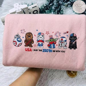 Op de afbeelding: Lichtroze sweatshirt met geborduurde Star Wars-personages in patriottische kleding. Het ontwerp omvat BB-8, Chewbacca, een Stormtrooper, Grogu, R2-D2 en Darth Vader, met de tekst "USA - May the 250th Be With You."