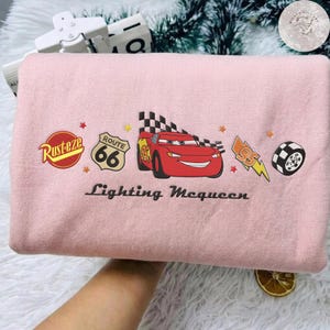 May include: A pale pink sweatshirt with a Lightning McQueen graphic from the Cars film. The design includes the Route 66 sign, the Rusteze logo, and the text "Lightning McQueen".