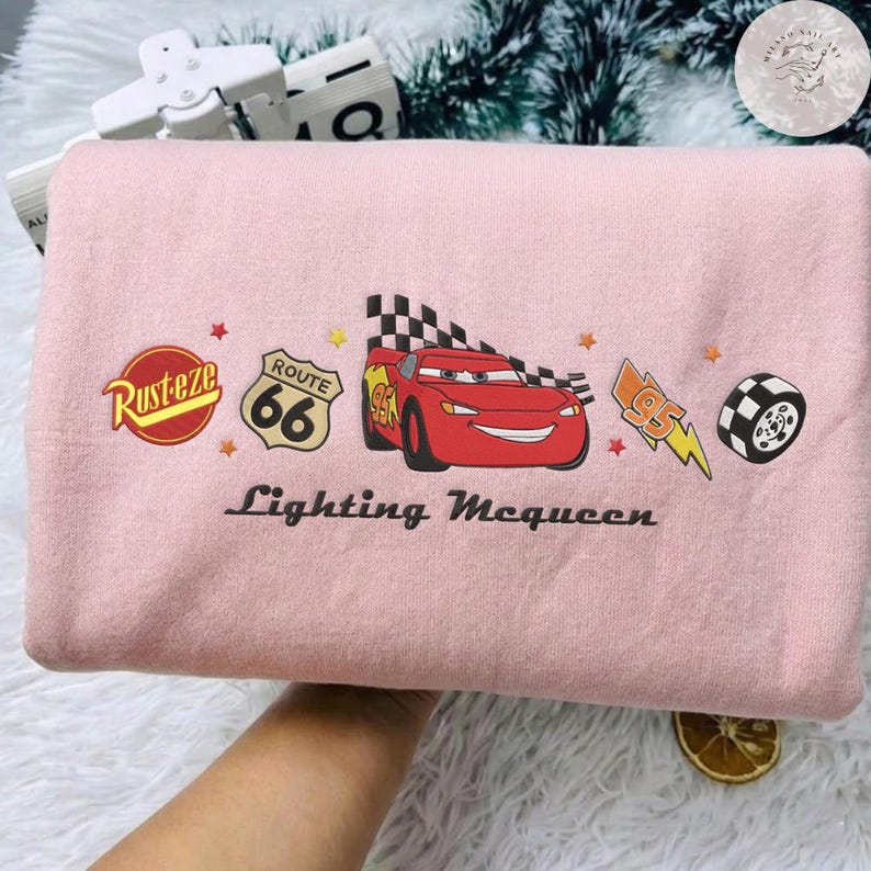 May include: A pale pink sweatshirt with a Lightning McQueen graphic from the Cars film. The design includes the Route 66 sign, the Rusteze logo, and the text "Lightning McQueen".