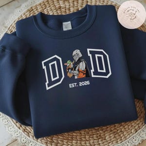 May include: Navy blue sweatshirt with a white outlined "DAD" graphic and an illustration of a character holding a small green creature. The text "EST. 2026" is below the illustration.