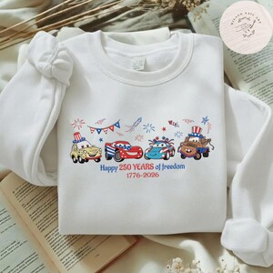 Pode incluir: Camisola branca com um gr&aacute;fico colorido de carros de desenho animado decorados com temas patri&oacute;ticos. Os carros s&atilde;o em vermelho, branco e azul, com fogos de artif&iacute;cio e o texto "Happy 250 Years of Freedom 1776-2026".