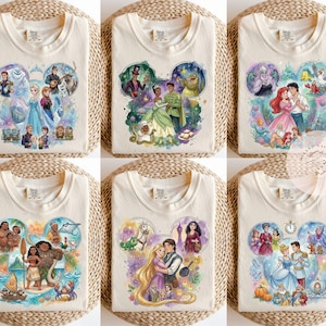 May include: A collection of six cream-colored t-shirts, each featuring a watercolor-style graphic of various animated characters arranged in a Mickey Mouse ear silhouette. The designs depict scenes from popular films including Frozen, The Princess and the Frog, The Little Mermaid, Moana, Tangled, and Cinderella.
