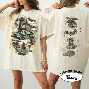 May include: Ivory-coloured t-shirt featuring graphics from The Mandalorian series. The front displays the text "The Mandalorian and Grogu" with character illustrations. The back has additional character and vehicle graphics. The shirt is labelled "Comfort Colors".