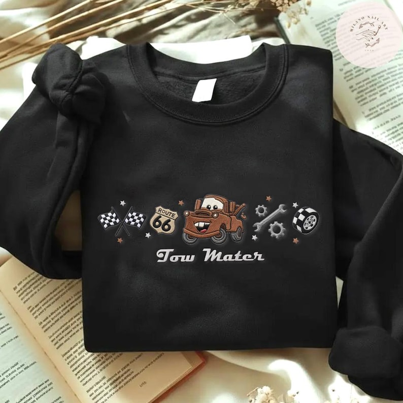 May include: A black long-sleeve sweatshirt featuring an embroidered graphic of a brown tow truck character, a Route 66 sign, checkered flags, a wrench, gears, and a tire. The text Tow Mater is embroidered in white below the central truck graphic.