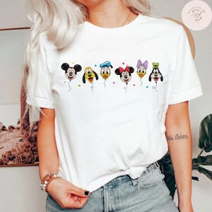 May include: White t-shirt featuring a colorful graphic of cartoon character balloons. The balloons depict Mickey Mouse, Pluto, Donald Duck, Minnie Mouse, Daisy Duck, and Goofy. The shirt has short sleeves and a crew neck.