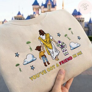 May include: Beige sweatshirt with a colourful graphic featuring Jesus, Woody, and Buzz Lightyear. The phrase "YOU'VE GOT A FRIEND IN ME" is printed in red and yellow. The design includes stars, clouds, and parachuters.
