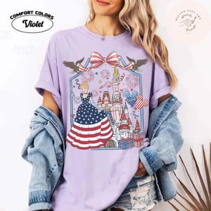 May include: A lilac Comfort Colors t-shirt with a patriotic Cinderella design. The graphic includes a castle, fireworks, and a dress with a stars and stripes pattern. The shirt has the text "Comfort Colors" and "Violet".