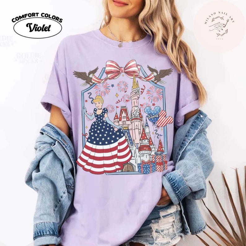 May include: A lilac Comfort Colors t-shirt with a patriotic Cinderella design. The graphic includes a castle, fireworks, and a dress with a stars and stripes pattern. The shirt has the text "Comfort Colors" and "Violet".
