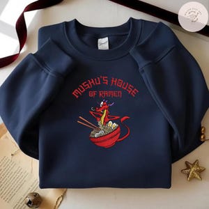 Op de afbeelding: Marineblauwe sweatshirt met een rode drakenillustratie die ramen eet uit een rode kom. De tekst "Mushu's House of Ramen" is in het rood geborduurd. De sweatshirt heeft een ronde hals en lange mouwen.