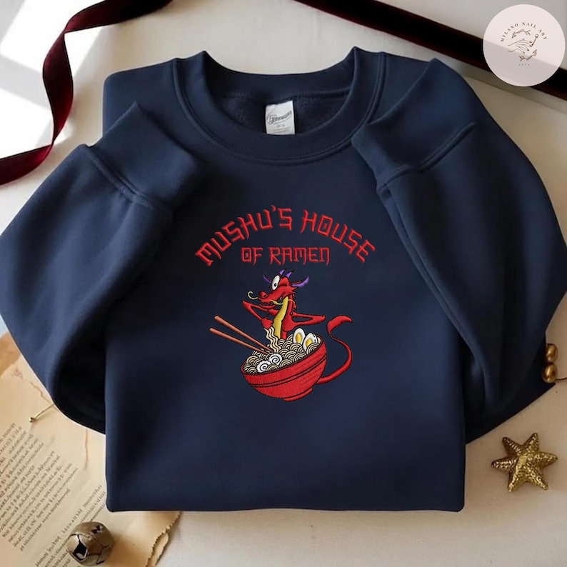 Op de afbeelding: Marineblauwe sweatshirt met een rode drakenillustratie die ramen eet uit een rode kom. De tekst "Mushu's House of Ramen" is in het rood geborduurd. De sweatshirt heeft een ronde hals en lange mouwen.