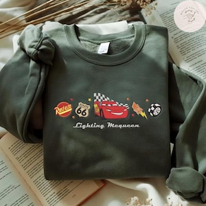 May include: An olive green sweatshirt with a graphic of Lightning McQueen from the film Cars. The design includes the Route 66 sign, a checkered flag, and the words "Lightning McQueen" in white text.