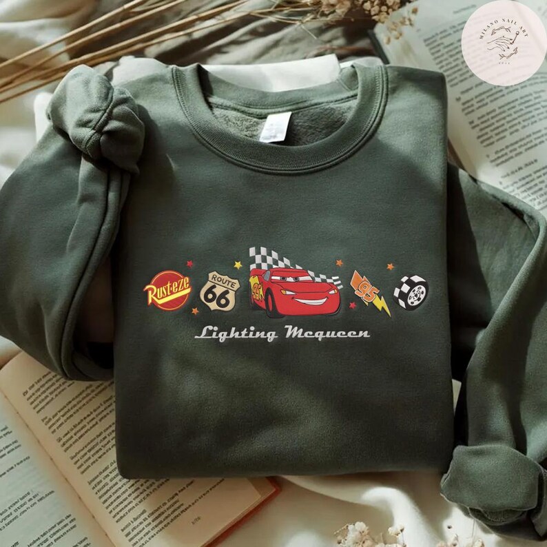 May include: An olive green sweatshirt with a graphic of Lightning McQueen from the film Cars. The design includes the Route 66 sign, a checkered flag, and the words "Lightning McQueen" in white text.