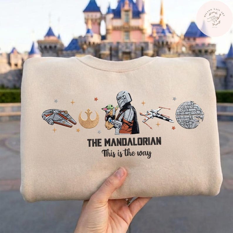 May include: Beige sweatshirt featuring embroidered images of the Millennium Falcon, a symbol, The Mandalorian with Grogu, an X-wing, and the Death Star. Text reads "THE MANDALORIAN" and "This is the way."