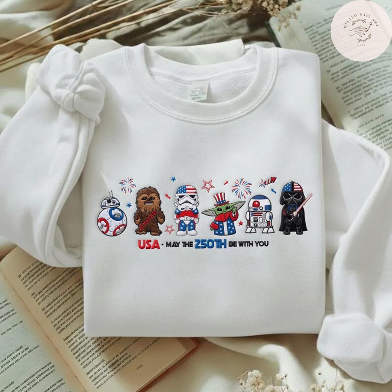 Op de afbeelding: Witte crewneck sweatshirt met geborduurde Star Wars-personages in patriottische outfits. Het ontwerp bevat BB-8, Chewbacca, een Stormtrooper, Grogu, R2-D2 en Darth Vader, met de tekst "USA - MAY THE 250TH BE WITH YOU."