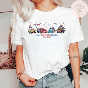 Pode incluir: Camiseta branca com um design patri&oacute;tico. Apresenta carros de desenho animado em vermelho, branco e azul, celebrando 250 anos de liberdade. O texto "Happy 250 YEARS of Freedom 1776-2026" est&aacute; impresso abaixo.