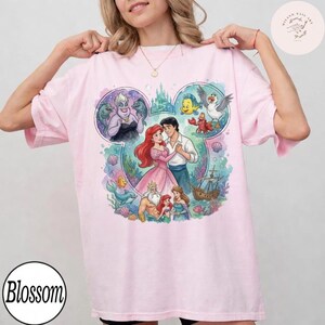 May include: A light pink t-shirt featuring a large graphic print of characters from The Little Mermaid arranged within a Mickey Mouse ear silhouette. The design includes Ariel, Prince Eric, King Triton, Ursula, Flounder, Sebastian, and Scuttle.