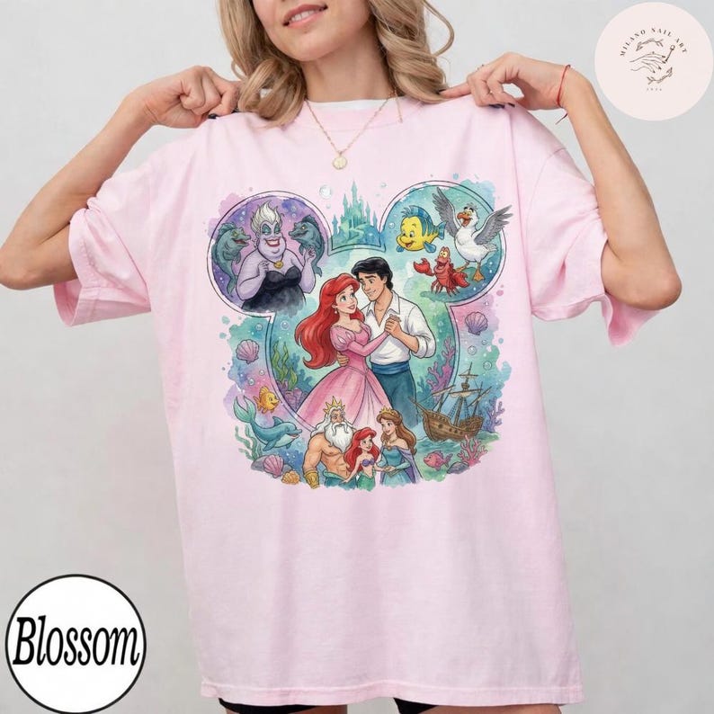 May include: A light pink t-shirt featuring a large graphic print of characters from The Little Mermaid arranged within a Mickey Mouse ear silhouette. The design includes Ariel, Prince Eric, King Triton, Ursula, Flounder, Sebastian, and Scuttle.