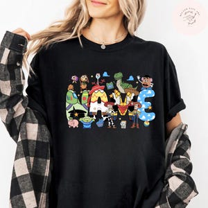 以下が含まれることがあります： 前面に大きなグラフィックプリントが施された黒の半袖Tシャツ。デザインには、トイ・ストーリーのキャラクターをモチーフにした模様で埋め尽くされた太字でカラフルな「NAME」という文字が描かれており、その周囲には様々なアニメキャラクターが配置されています。