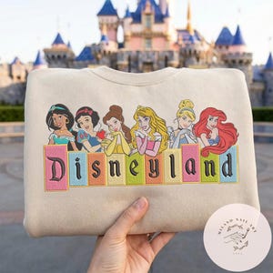 May include: Cream-coloured sweatshirt with embroidered Disney princesses and the word "Disneyland" in colourful letters. The princesses include Jasmine, Snow White, Belle, Aurora, Cinderella, and Ariel. A castle is in the background.