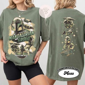 May include: Olive green t-shirt with a graphic design from the Star Wars series, "The Mandalorian and Grogu." The front of the shirt displays the title and characters. The back of the shirt has additional character graphics. The shirt is labelled "Comfort Colors Moss."