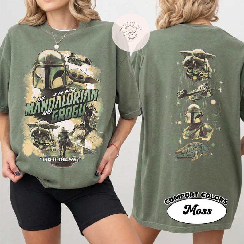 May include: Olive green t-shirt with a graphic design from the Star Wars series, "The Mandalorian and Grogu." The front of the shirt displays the title and characters. The back of the shirt has additional character graphics. The shirt is labelled "Comfort Colors Moss."