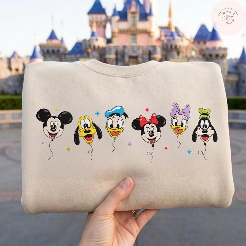 May include: Beige crewneck sweatshirt featuring embroidered Disney characters as balloons. Mickey Mouse, Pluto, Donald Duck, Minnie Mouse, Daisy Duck, and Goofy are depicted with smiling faces and colorful accents. A fun, whimsical design.