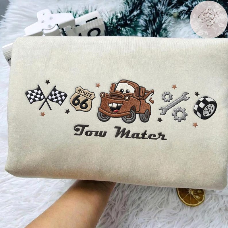 May include: A cream-colored fabric item featuring an embroidered brown tow truck character, a Route 66 sign, two checkered flags, a wrench, gears, and a tire. The text Tow Mater is embroidered in dark brown below the central truck graphic.