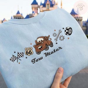 May include: A light blue crewneck sweatshirt featuring embroidered designs including a brown tow truck, a Route 66 sign, two checkered flags, a wrench, gears, and a tire. The text Tow Mater is embroidered in black script below the truck graphic.