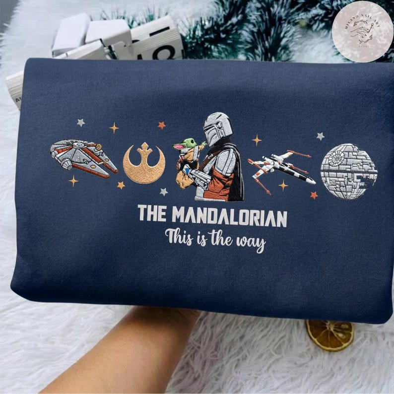 May include: Navy blue blanket featuring embroidered images of the Mandalorian, Baby Yoda, and Star Wars spacecraft. The text "THE MANDALORIAN This is the way" is printed below the images. The blanket is folded.